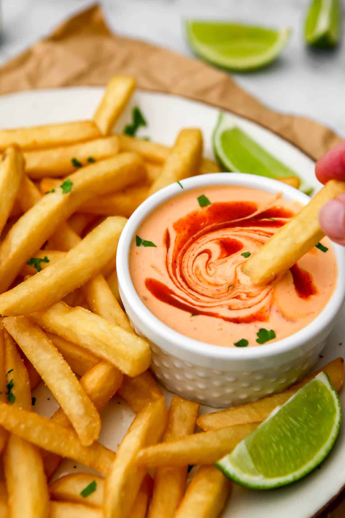 French fries on a white plate being dipped into a small white bowl of vegan sriracha mayo with slices of limes around it.