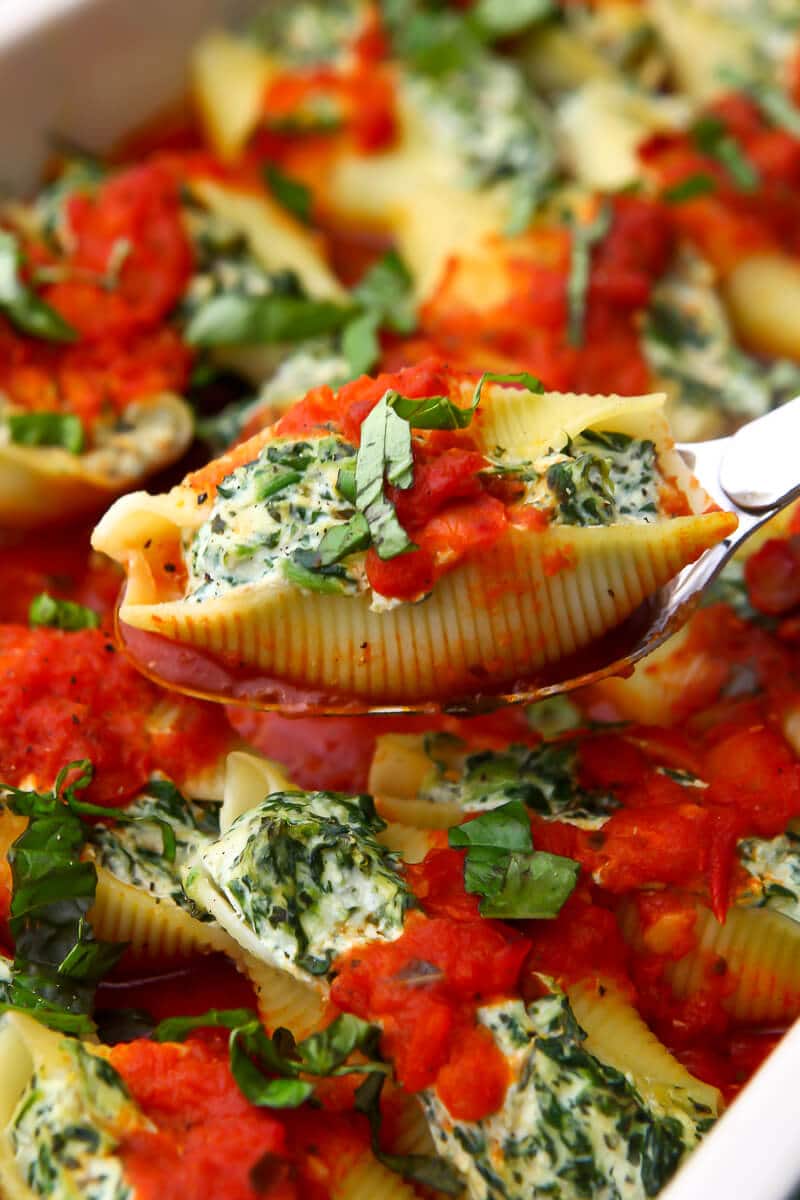 A spoon lifting a jumbo stuffed shell stuffed with tofu ricotta and spinach out of a baking dish.