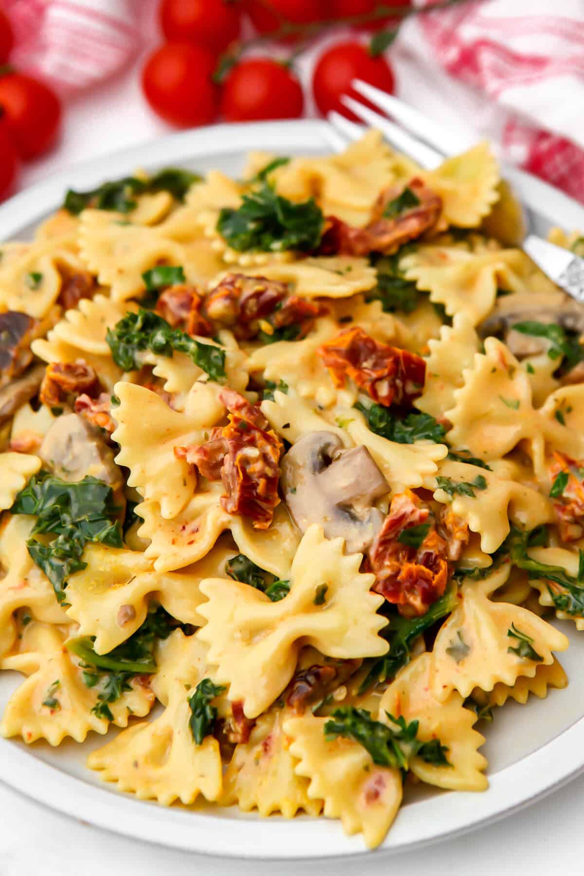 A white plate with vegan sundried tomato pasta with cherry tomatoes in the background.