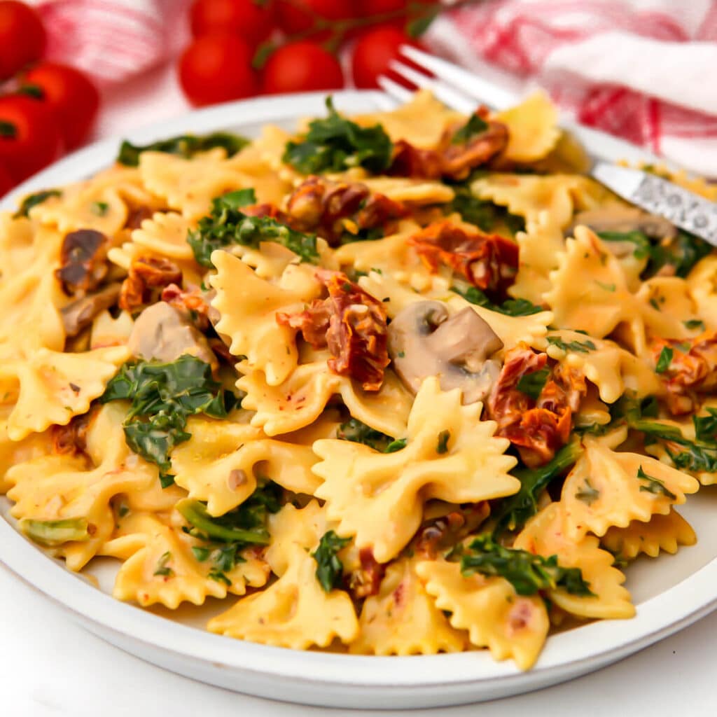 A serving of creamy vegan sundried tomato pasta with mushrooms and kale on a white plate.