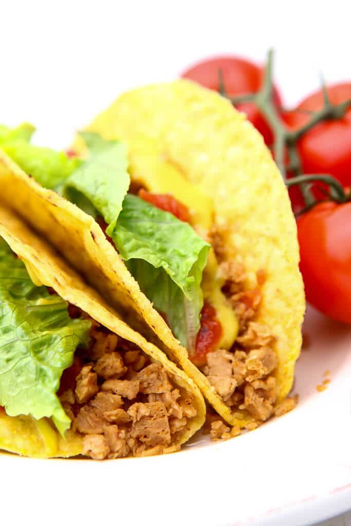 Veggie tacos made with TVP taco meat on a white plate with tomatoes on the side.