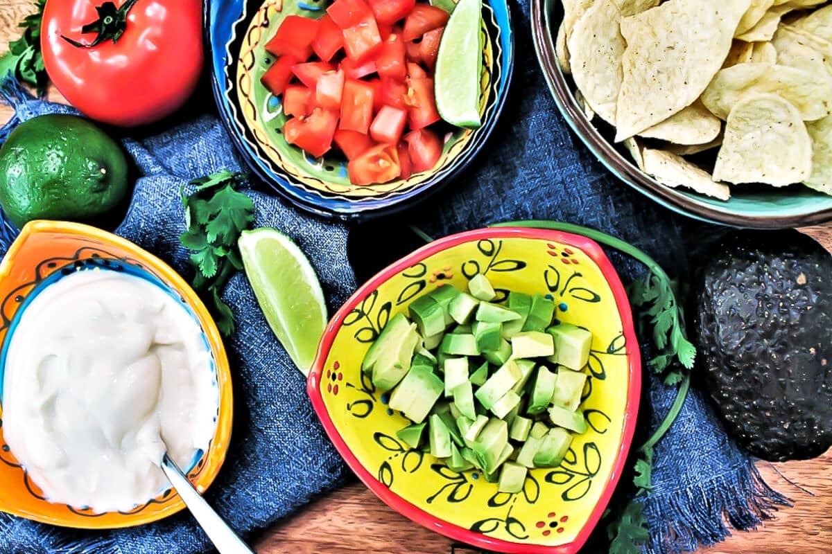 A toppings bar for vegan taco soup with vegan sour cream, avocado, salsa, and corn chips.