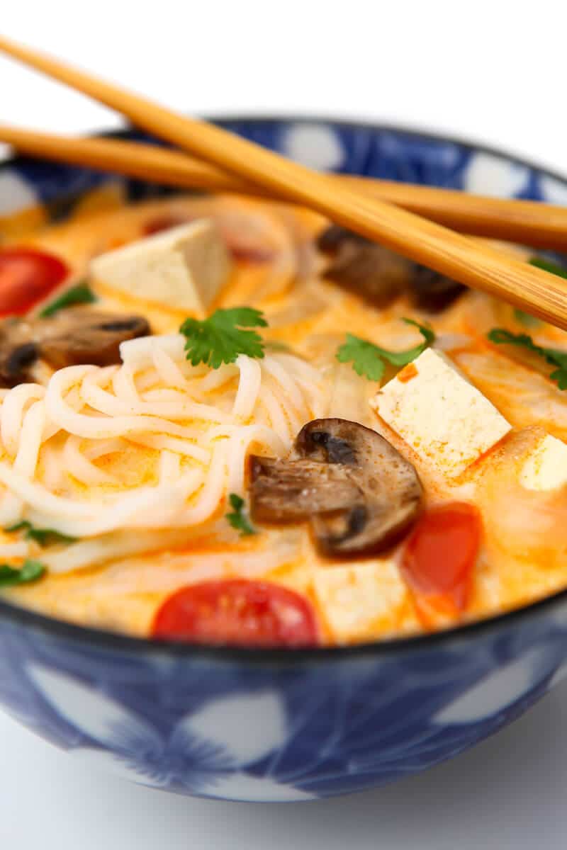 A bowl of vegetarian tom kha soup with veggies, tofu, and herbs with chopsticks on top.