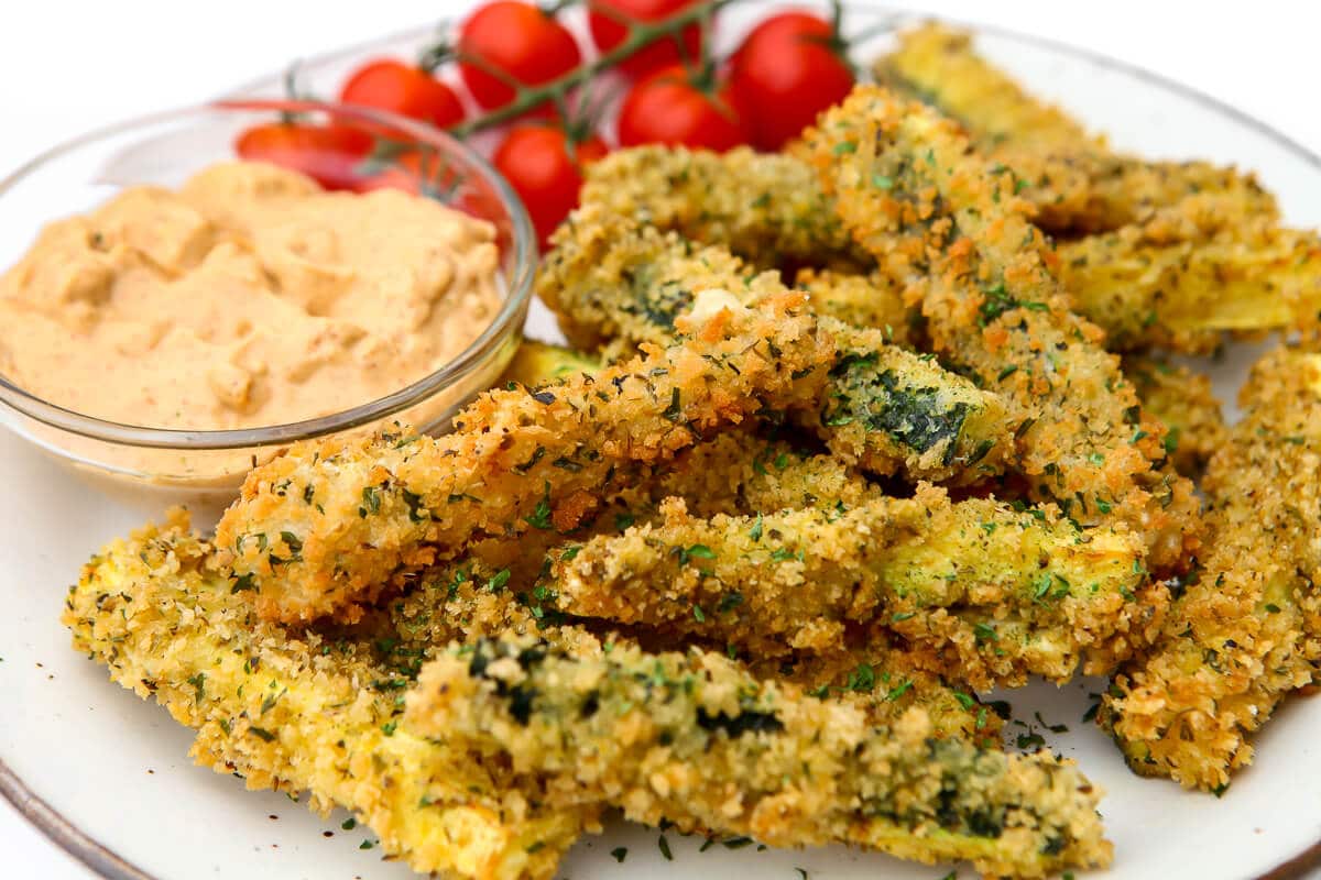 A white plate filled with breaded deep fried zucchini cut in to long strips like fries with chipotle dipping sauce on the side.