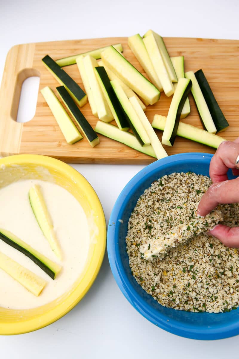 Cut zucchini on a cutting board with a bowl of batter and a bowl of bread crumbs to coat the zucchini in before frying.