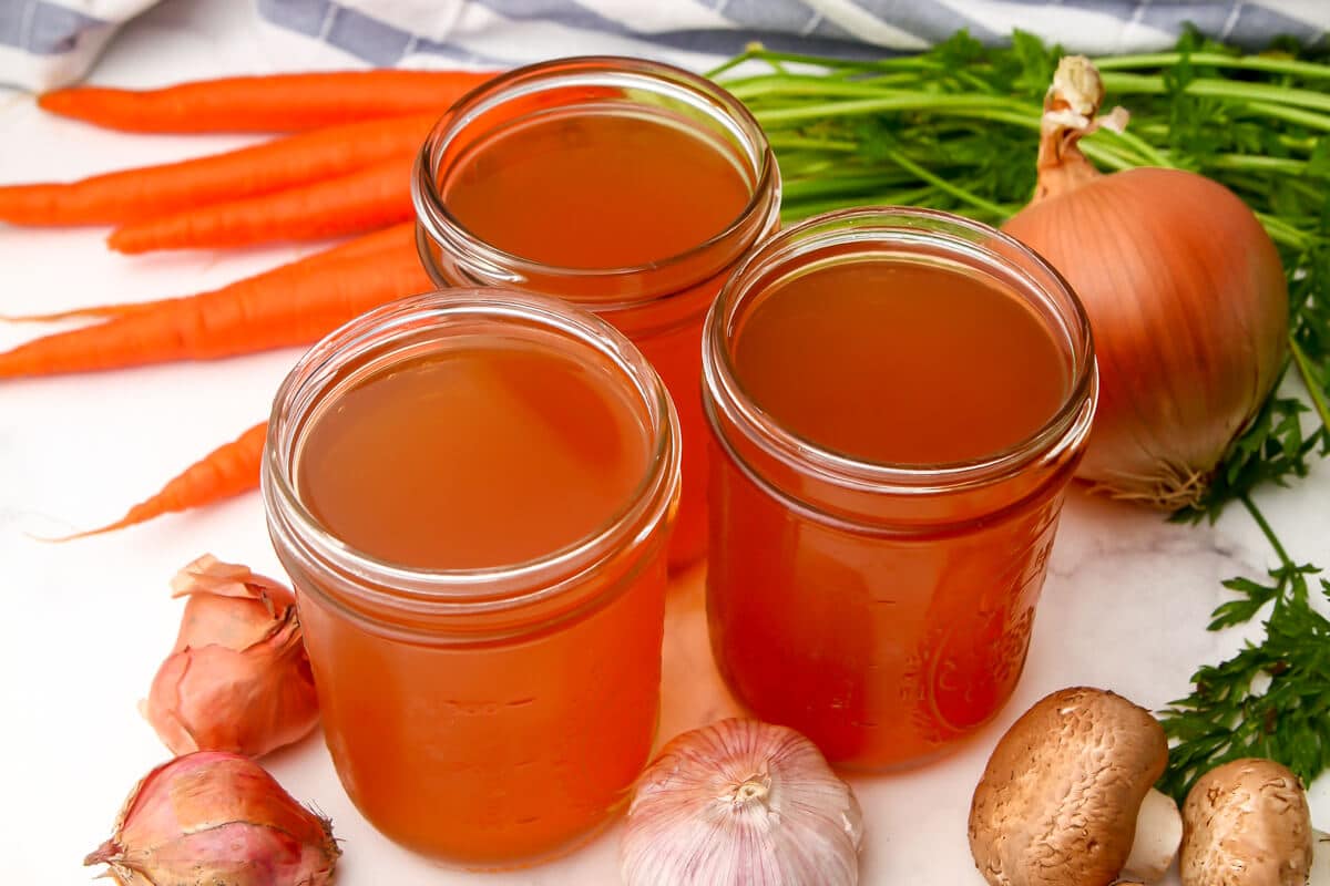 Vegan broth in jars next to vegetables with a tea towel behind it.