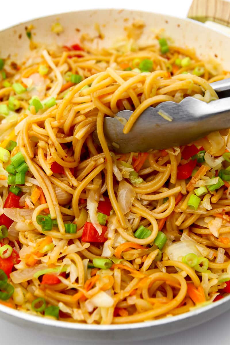 Noodles being tossed with cooked veggies and spicy oil sauce topped with green onions.